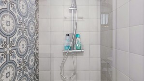 Een douche met een handdouche en een wandmengkraan, een plank met toiletartikelen en getextureerde tegels op de muur.
