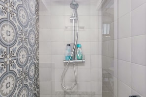 A shower with a handheld showerhead and a wall-mounted faucet, a shelf with toiletries, and patterned tiles on the wall.