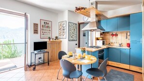 A modern kitchen with blue cabinets, a dining area with a round table and chairs, a TV on a cart, and a balcony with a view.