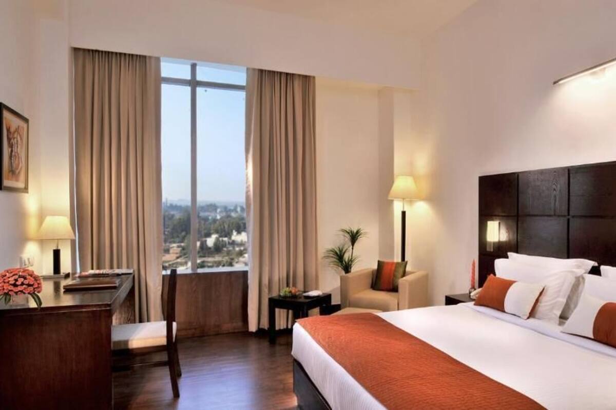 Ein Hotelzimmer mit einem großen Bett, einem Schreibtisch, einem Stuhl, einem kleinen Tisch und Blick auf die Stadt durch große Fenster.