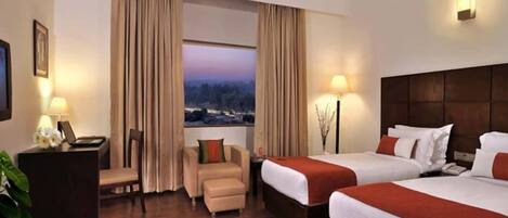 A hotel room with two beds, a desk, a chair, and a view of a cityscape.