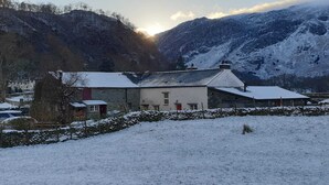 Eine verschneite ländliche Landschaft mit einem Steinhaus, das ein geneigtes Dach und ein kleines Angebautes hat.