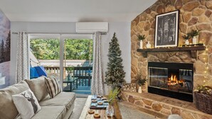 Ein gemütliches Wohnzimmer mit Kamin aus Naturstein, einer Couch, einem Couchtisch mit Dekoration und Blick ins Freie durch eine Schiebetür.