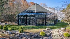 Ein moderner Glasanbau mit einem Steinpfad, umgeben von einem gepflegten Garten und einem bewaldeten Gebiet.
