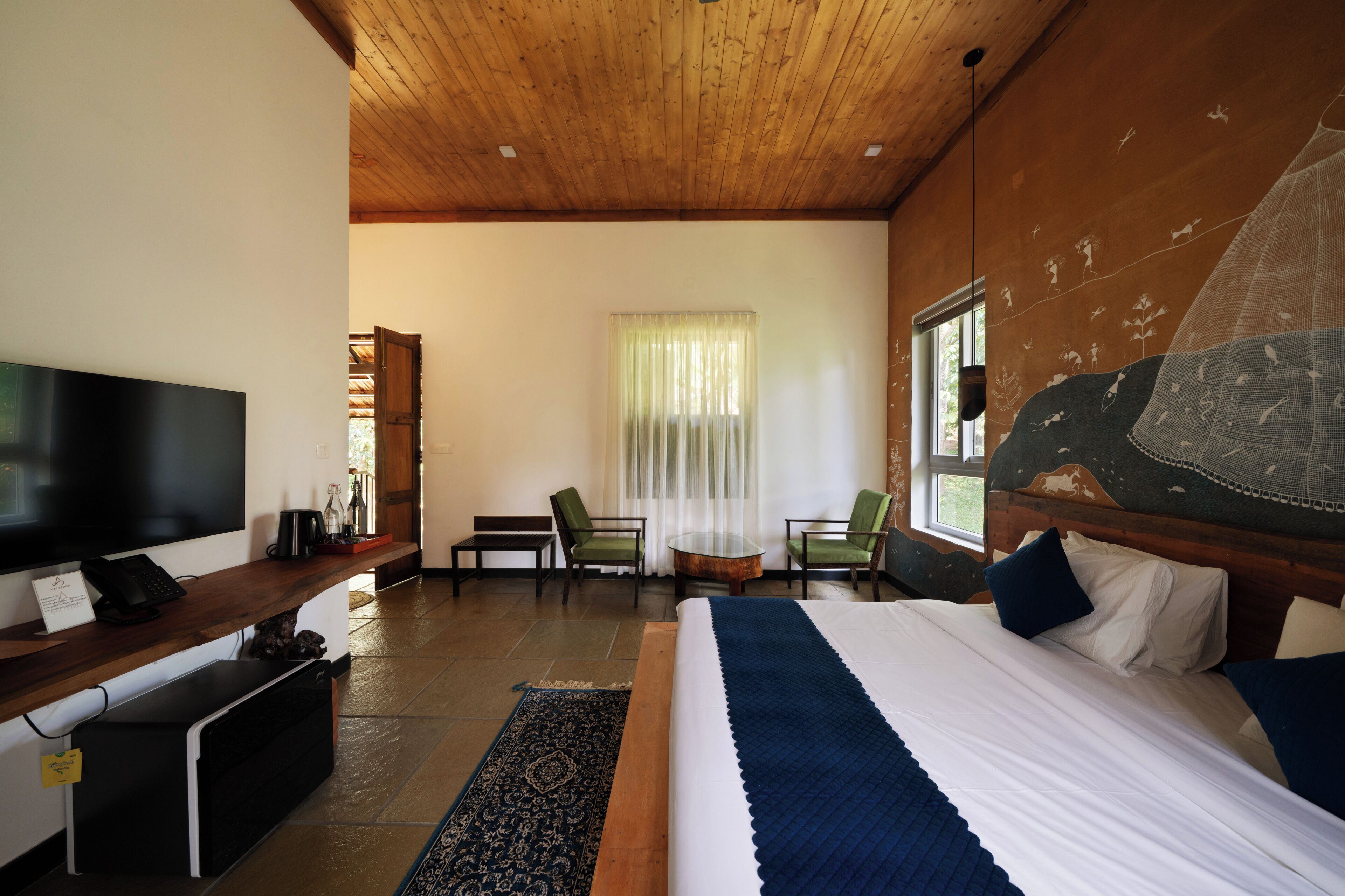 Habitación de hotel con cama, escritorio con computadora, televisor y ventana con vista a follaje.