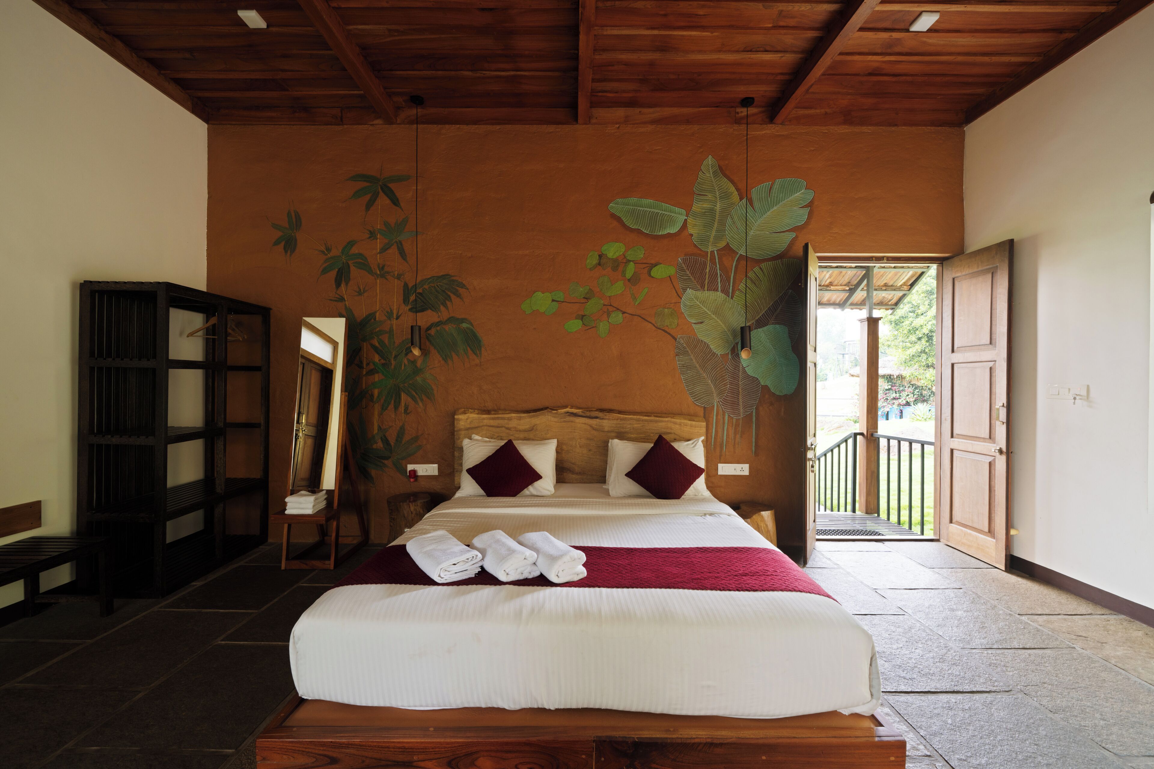 Une chambre avec une tête de lit en bois, un lit avec une literie blanche, un plaid rouge et un grand miroir. On y trouve également une armoire en bois et une porte donnant sur l’extérieur.