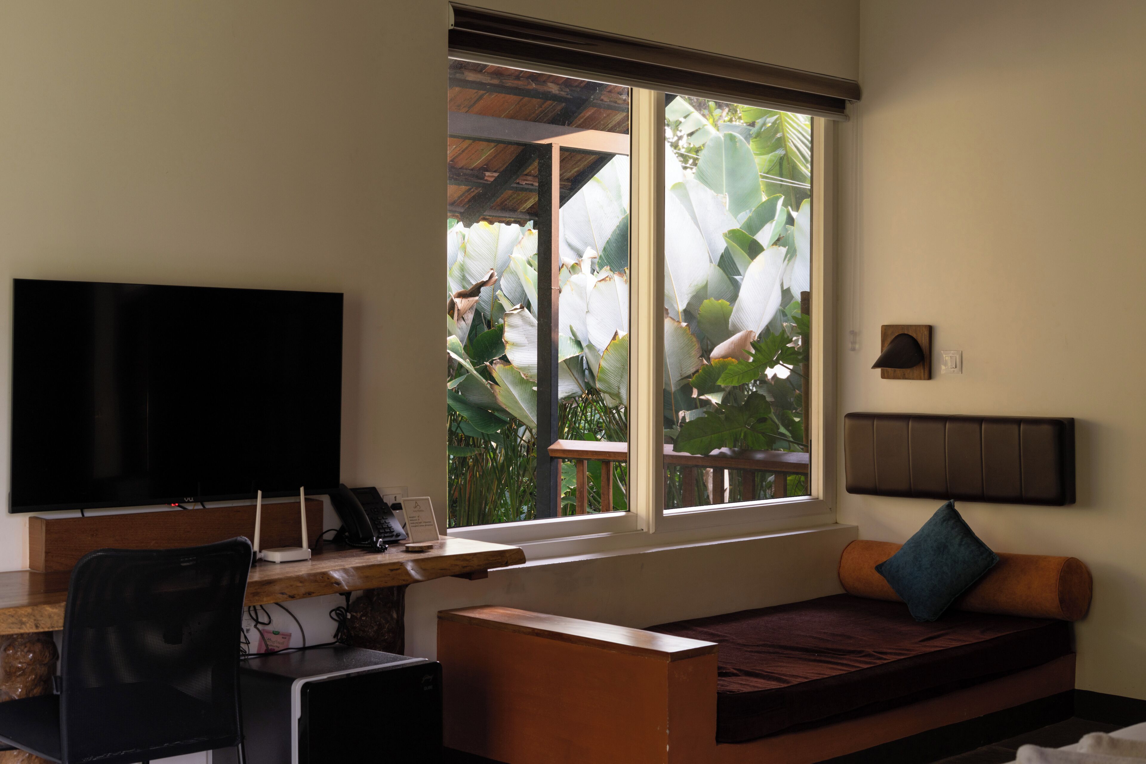 A room with a television, a desk with a computer, a sofa, and a window with a view of plants.