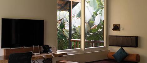 A room with a television, a desk with a computer, a sofa, and a window with a view of plants.