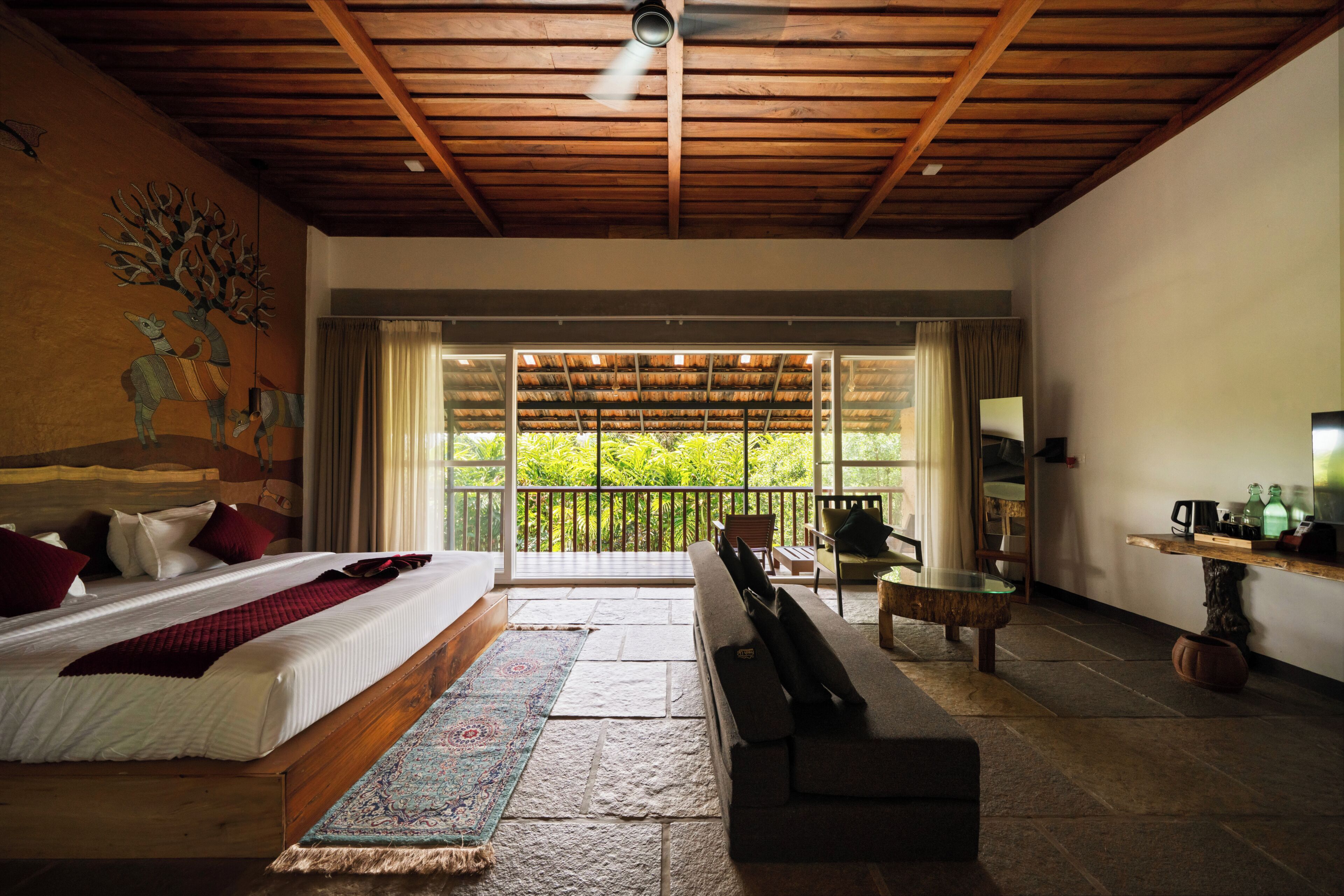 Amplia habitación con una cama grande, un banco, un escritorio y un balcón con vistas a zonas verdes.
