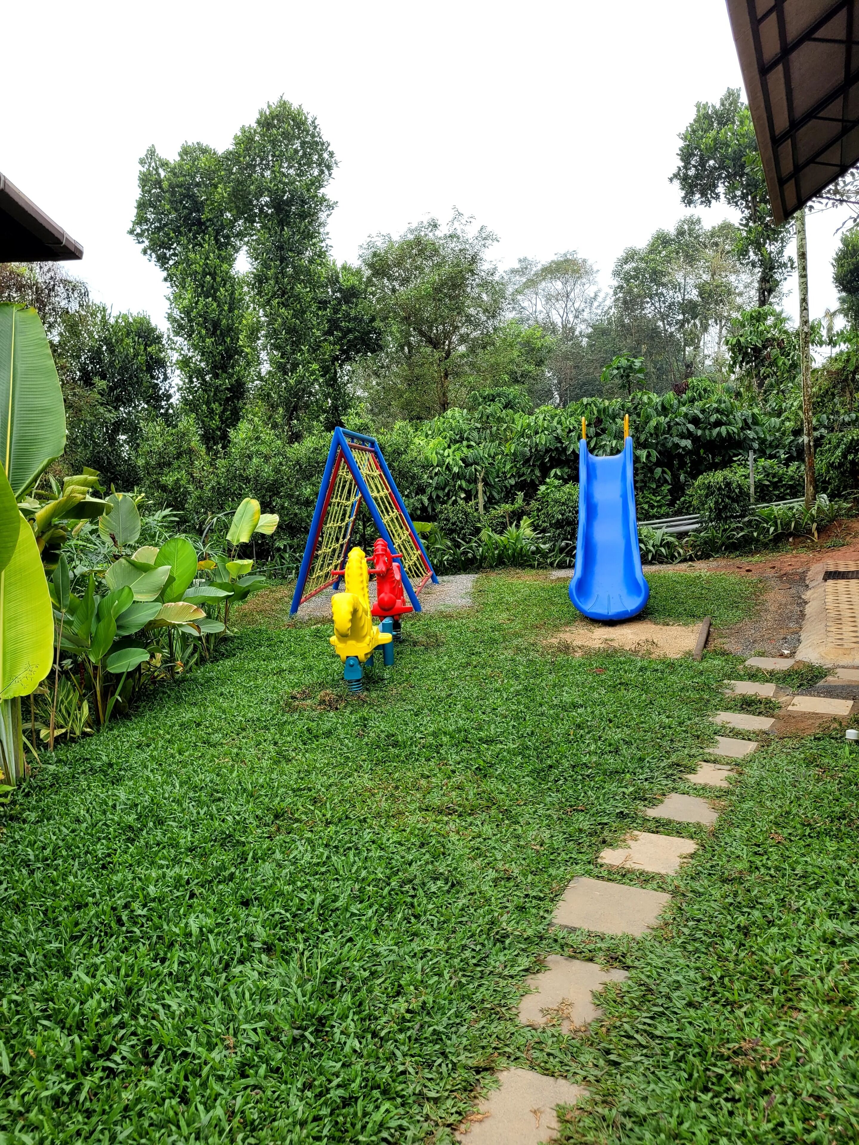 A playground with a climbing structure and a slide, surrounded by lush greenery and a paved path.