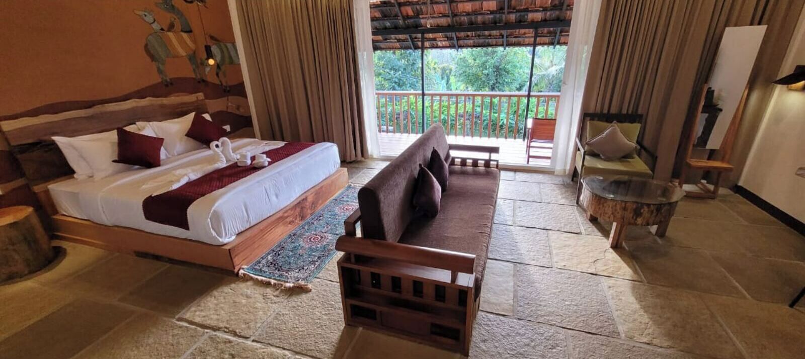 A hotel room with a bed, a sofa, a small table, and a balcony with a view of greenery.
