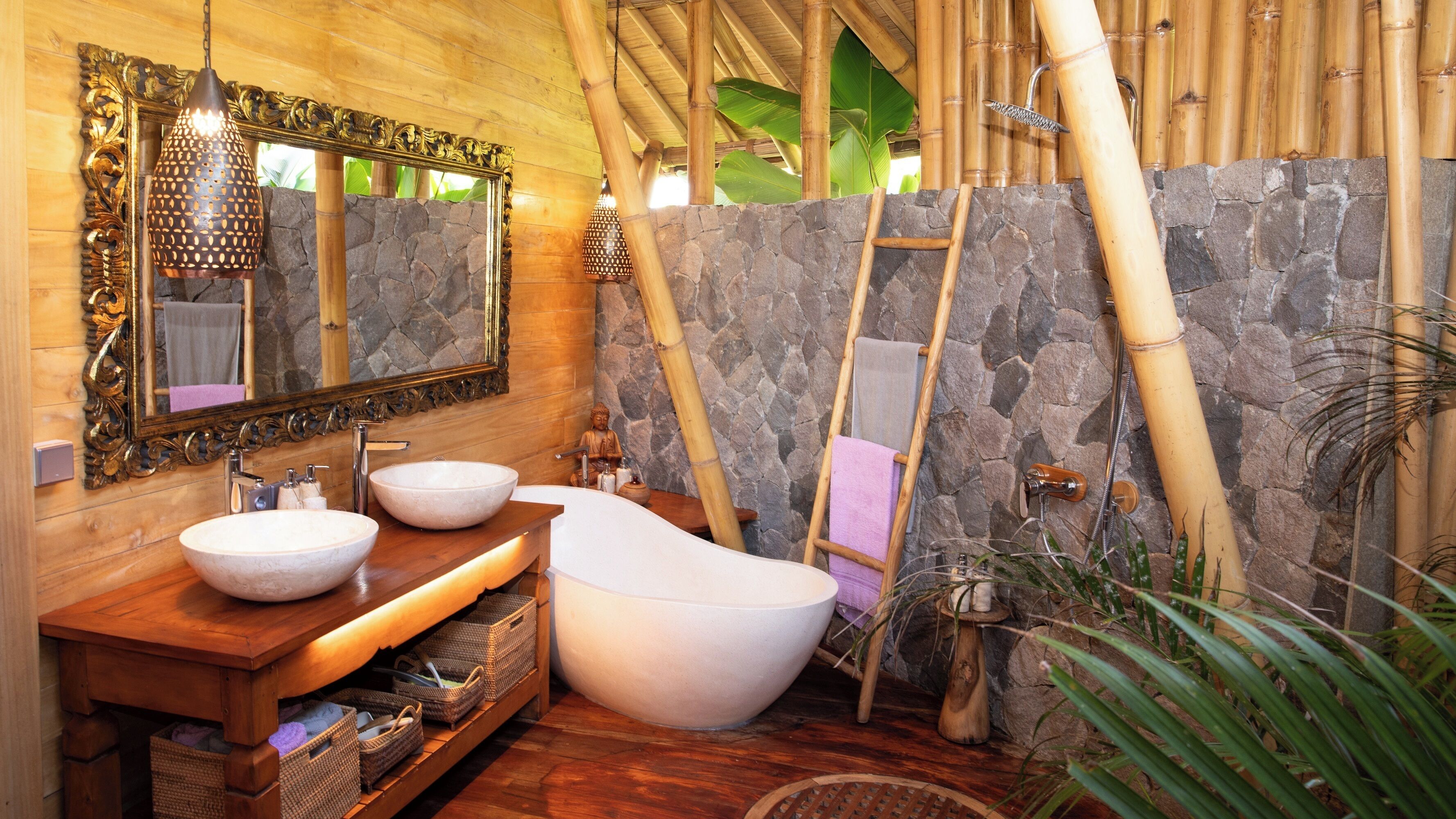 A bathroom with a wooden vanity, two sinks, a freestanding bathtub, a stone wall, a bamboo ladder, and a large mirror.