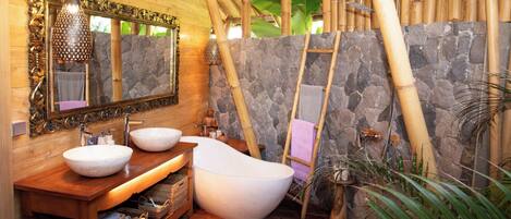A bathroom with a wooden vanity, two sinks, a freestanding bathtub, a stone wall, a bamboo ladder, and a large mirror.