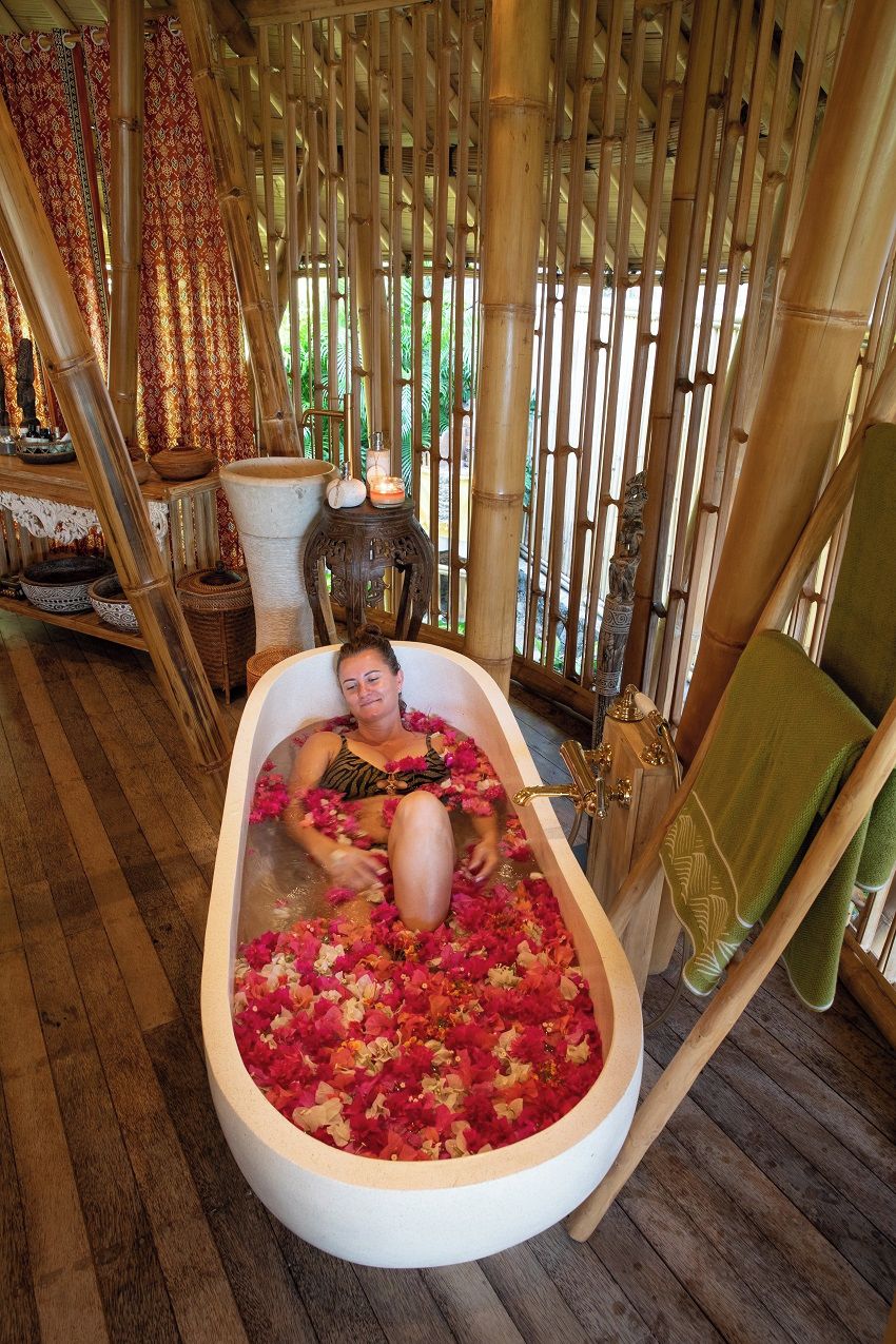 A person relaxing in a bathtub filled with rose petals, surrounded by bamboo decor and traditional furnishings.