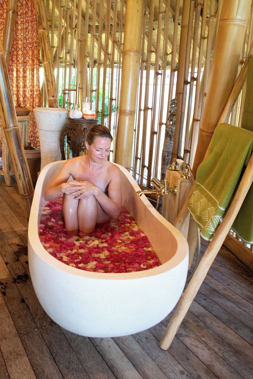 A person in a bathtub filled with rose petals, surrounded by bamboo structures and a green towel on a chair.