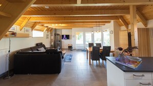 Amplia sala de estar con techo de madera, un sofá negro, una mesa de comedor con sillas y una zona de cocina con fregadero y estufa.