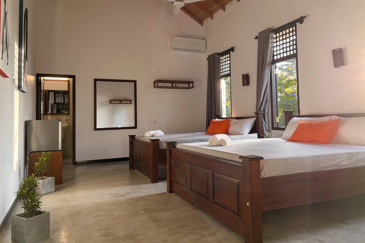 A bedroom with two beds, wooden headboards, a ceiling fan, an air conditioning unit, and a view of greenery outside.