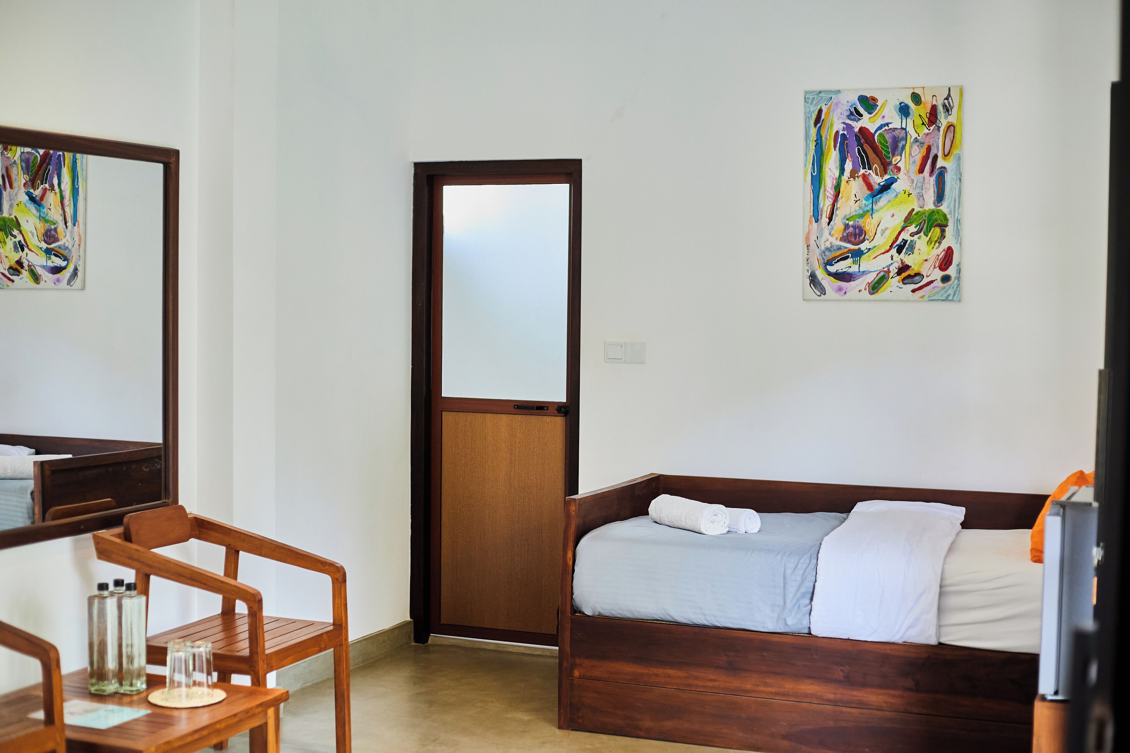 A room with a wooden bed, a wooden chair, a small table with a glass, and a painting on the wall.