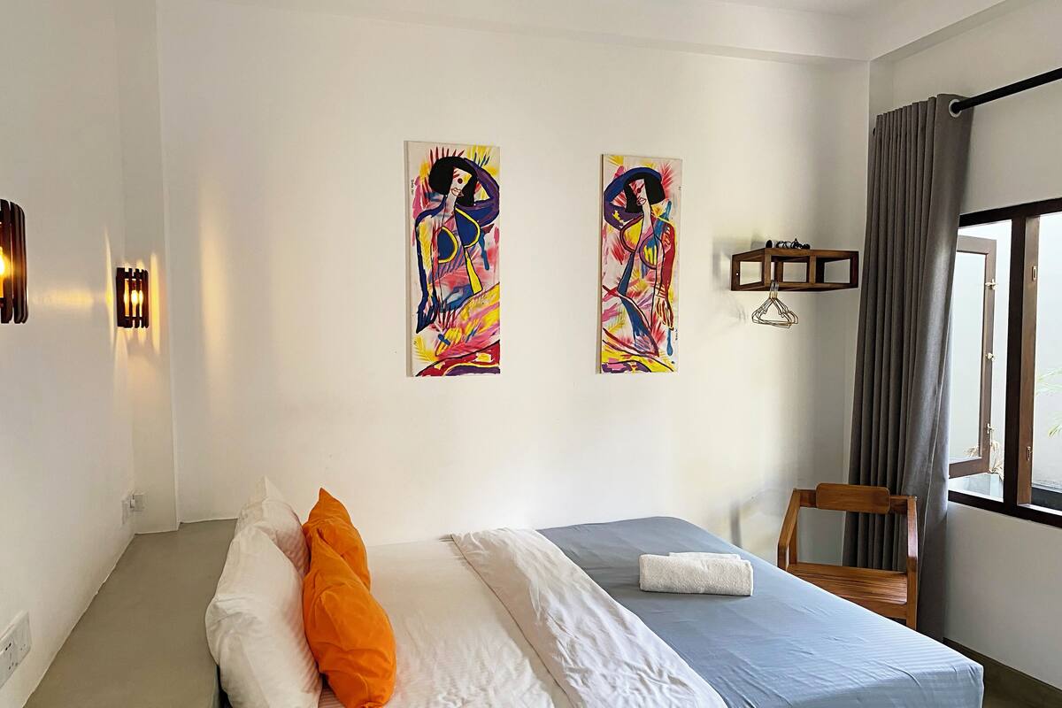 A hotel room with a bed, a chair, and two abstract paintings on the wall.
