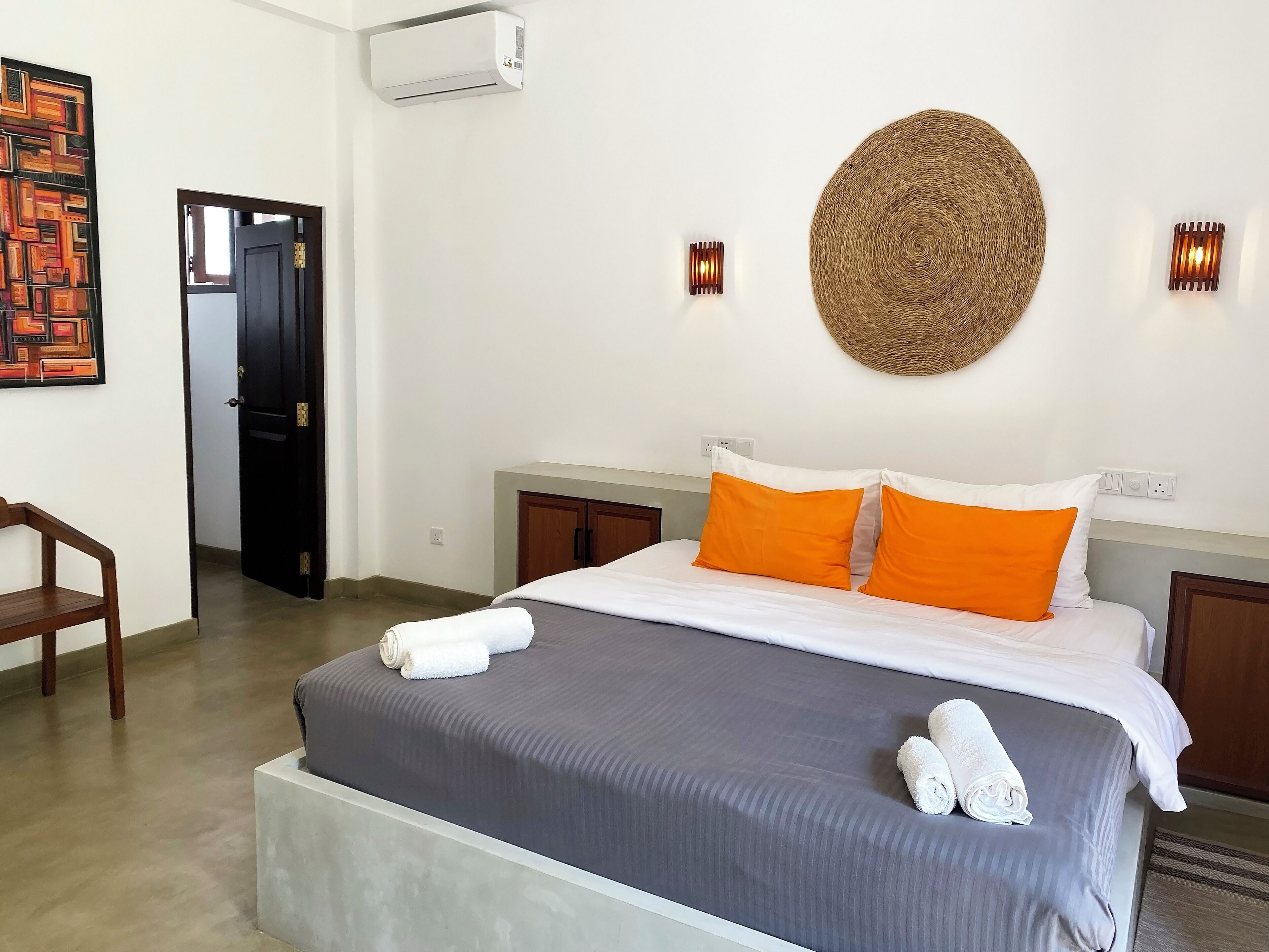 Habitación de hotel con una cama, dos mesitas de noche, aire acondicionado de pared, una puerta y una obra de arte en la pared.