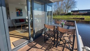 Een modern terras buiten met een glazen deur naar een leefruimte, een tafel met stoelen en uitzicht op een meer en bomen.