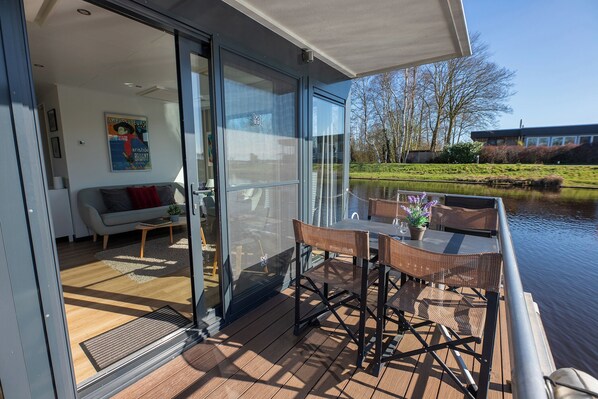 Una terraza moderna al aire libre con puerta de vidrio que da a un salón, una mesa con sillas y vistas a un lago y árboles.
