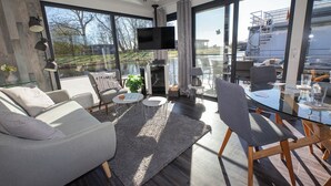 Un salon moderne comprenant un canapé, une table basse et des fauteuils, une cheminée et de grandes fenêtres donnant sur un plan d’eau et un bâtiment.