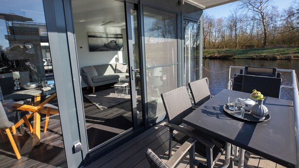 Un espace de repas extérieur moderne, avec une table dressée pour deux personnes, une pièce fermée par des baies vitrées et dotée d’un canapé, et une vue sur une rivière.