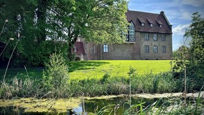 Un grand bâtiment en briques, doté de nombreuses fenêtres et d’un toit en pente, entouré d’une végétation luxuriante et d’un petit étang.