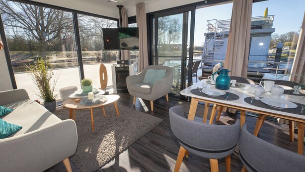 A modern living room with a dining area, large windows, and a flat-screen TV. - Houseboat 7 Starfish. Picturesque by the lake and unconventional. (Geestland)
