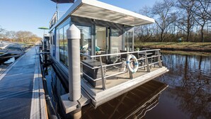 A modern houseboat with a covered deck, glass walls, and a lifebuoy. - Houseboat 7 Starfish. Picturesque by the lake and unconventional. (Geestland)