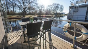 A deck with a table and chairs, a lifebuoy, and a view of a river and trees. - Houseboat 7 Starfish. Picturesque by the lake and unconventional. (Geestland)