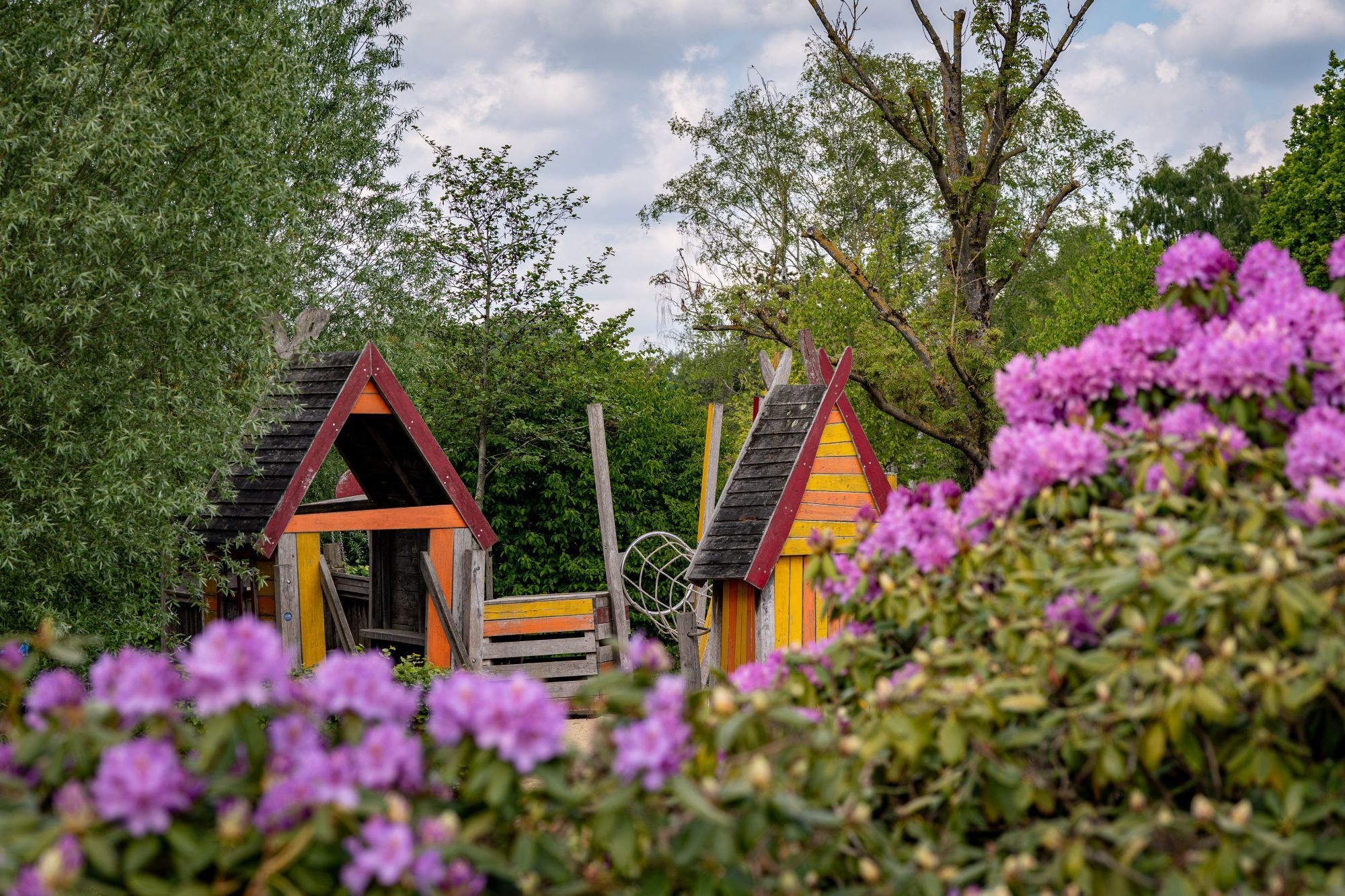 Two wooden playhouses with triangular roofs, surrounded by lush greenery and purple flowers.