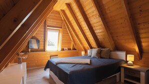 Ein Schlafzimmer mit schrägem Dach, Holzwänden, einem Bett mit dunkler Tagesdecke, einer Nachttischlampe und einem Fenster mit Blick.