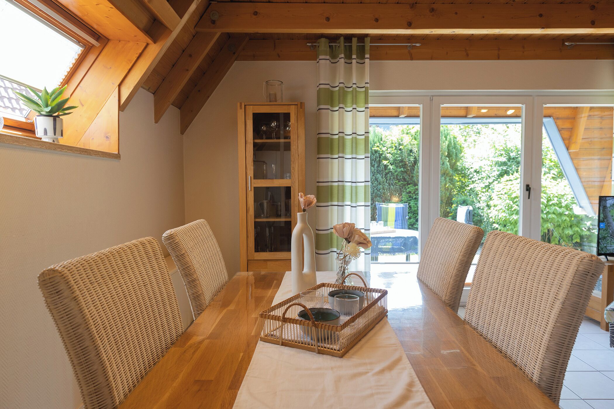 A dining area with a wooden table, wicker chairs, a glass cabinet, and a view of greenery outside.