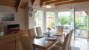 A dining area with a wooden table set for four, surrounded by chairs, with a view of the garden through large windows. - Vacation home Westertill 15 with tidal magic and at the cutter harbor. (Wurster Nordseeküste)