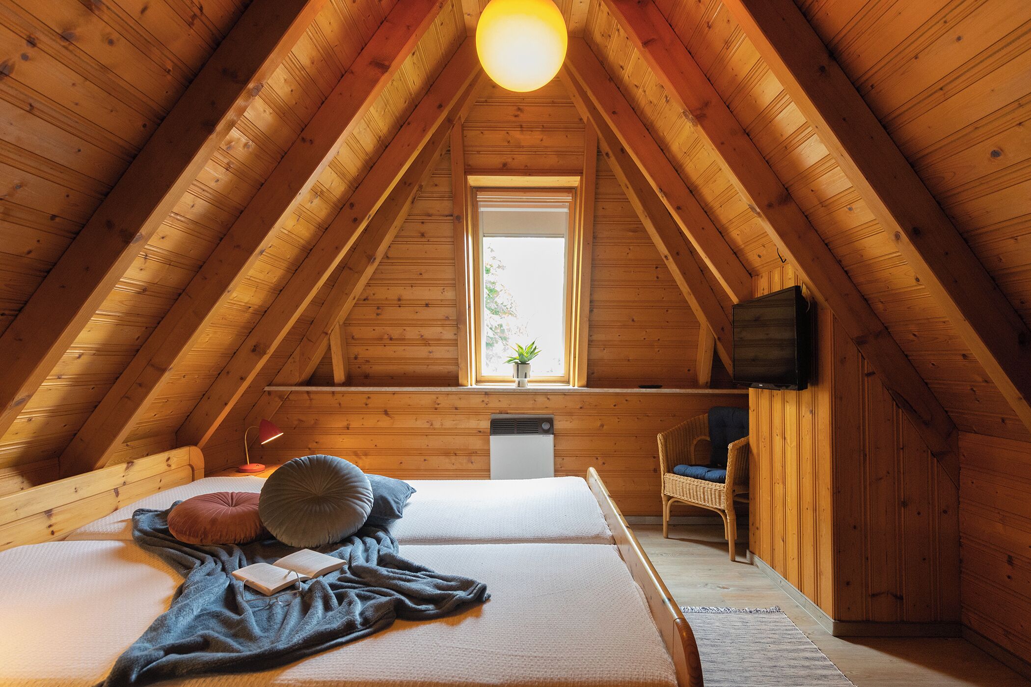 Un dormitorio acogedor en el ático, con techo inclinado, cama con almohadas y manta, una silla, una mesita y una ventana con una planta.