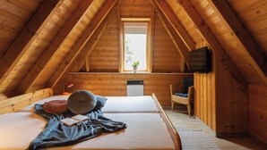 A cozy attic bedroom with a sloped ceiling, a bed with pillows and a blanket, a chair, a small table, and a window with a plant.