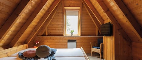 A cozy attic bedroom with a sloped ceiling, a bed with pillows and a blanket, a chair, a small table, and a window with a plant.