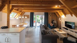A modern loft-style living space with a high ceiling, wooden beams, a dining area, a sofa, a TV, and a fireplace.