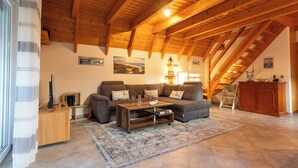 A spacious living room with a high wooden ceiling, a brown sofa set, a wooden coffee table, and a large rug. - Vacation home Piratennest. Child-friendly and close to the beach. (Wurster Nordseeküste)