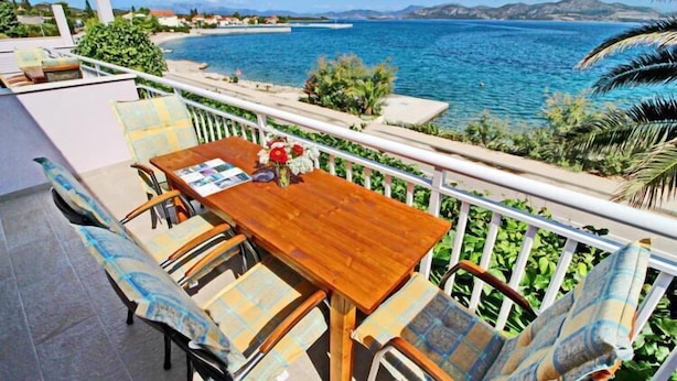 A balcony with a wooden table and chairs, overlooking a coastal area with a view of the sea and distant mountains.