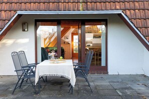 Ein Essbereich im Freien mit Tisch und Stühlen, einer weißen Tischdecke und Blick ins Haus durch eine Glastür.