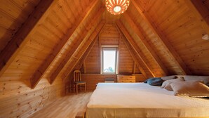 Quarto de sótão acolhedor com teto inclinado de madeira, uma cama de solteiro, uma cadeira e uma pequena janela.