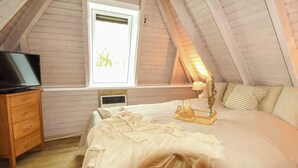 Un dormitorio con techo inclinado, un televisor sobre una cómoda de madera, una cama con colcha blanca y una ventana con vista a árboles.