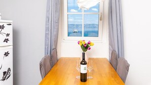 Une table à manger avec une bouteille de vin et des verres, un vase rempli de fleurs et une vue sur la mer par une fenêtre ornée de rideaux.
