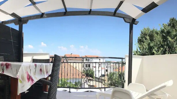 Uma varanda com toalha de mesa branca, uma cadeira e vista para edifícios e árvores.