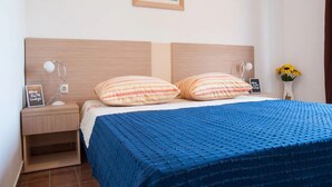 Cama bem arrumada com colcha azul texturizada e almofadas listradas, mesa de cabeceira com abajur, quadro na parede e um vaso com girassóis.