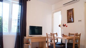 Quarto com televisão sobre uma mesa de madeira, uma mesa de jantar com cadeiras, uma janela com cortinas e um quadro pendurado na parede.