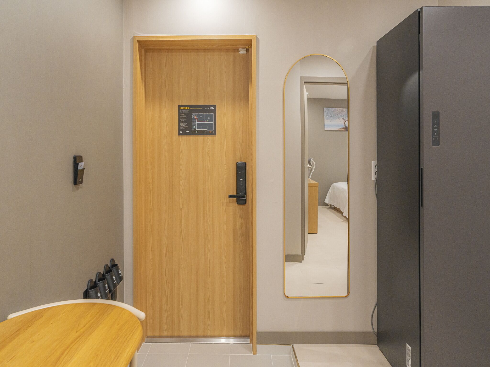 A hotel room with a wooden door, a mirror, a gray refrigerator, and a wooden bench.